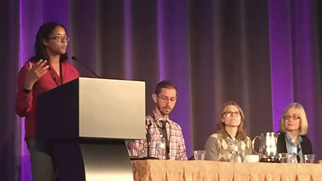 A woman speaking at a podium, and three individuals sitting at a panelist table at a conference
